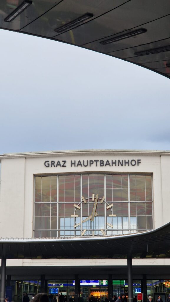 Graz Hauptbahnhof mit großer Uhr über dem Eingang, umgeben von moderner Dachkonstruktion und Glasfassaden.