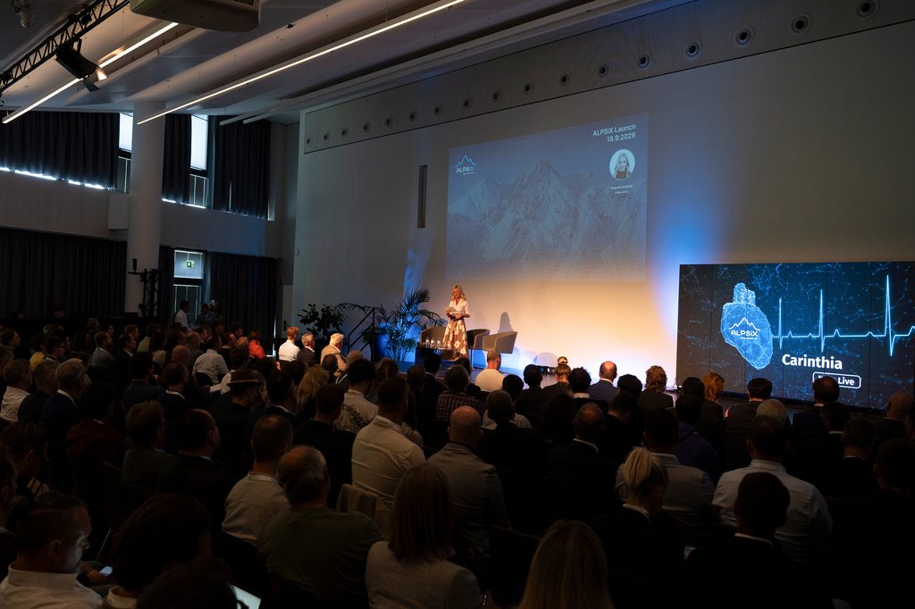 Eine Person hält einen Vortrag auf einer Bühne vor einem Publikum, mit Präsentation über ALPSIX Launch.