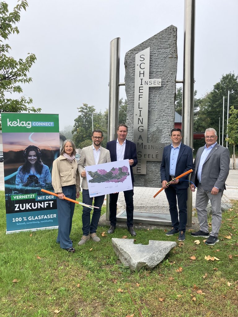 Fünf Personen vor einem Denkmal in Schiefling, Kärnten, halten ein Glasfaserkabel und eine Landkarte.